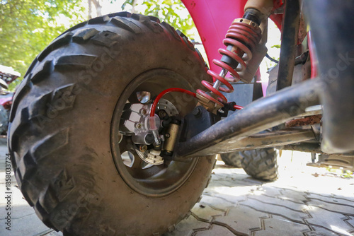 Detailed close up shot of dirty heavy duty black tire, strong red suspension spring and chassis of adventure off road ATV vehicle parked pavement outdoor showing tough vibe ready for extreme