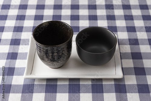 Two different black ceramic cups on white rectangular plate over blue checkered tablecloth