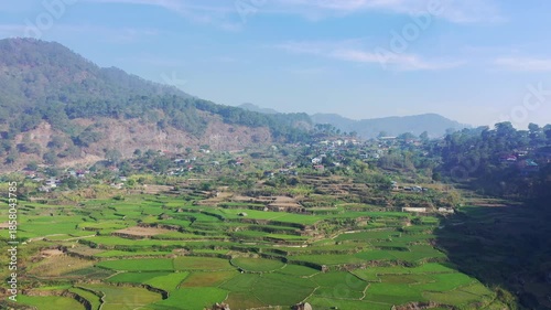 Aerial view of vibrant green rice terraces nestled in a mountainous landscape near Sagada, Philippines, with a rural village in the background under a clear blue sky.