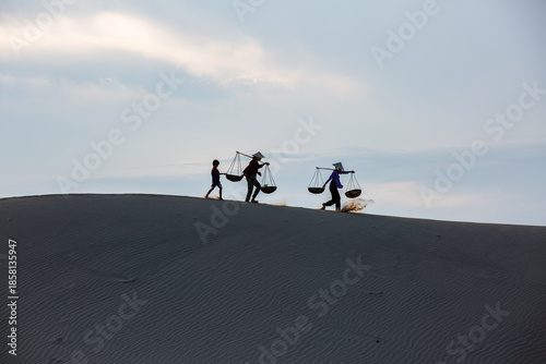 Cham women walking home across the sand dunes at sunset, beneath a blue sky filled with soft white clouds.