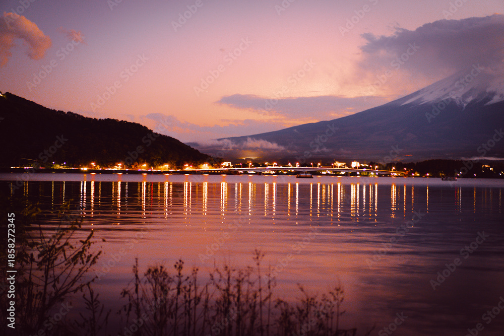 Obraz premium Night view of Mount Fuji with snow covering the peaks on a cloudy day at night. There are bridges over the lake and bokeh of colorful lights. There is reflection of water in the landscape.