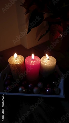 Three festive candles—gold, pink, and silver—burn brightly on a tray with Christmas ornaments. The warm flames create a cozy, magical atmosphere against a dark background with holiday greenery.