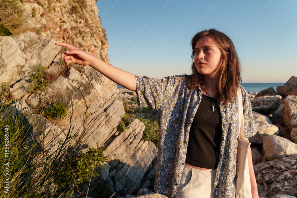 Naklejka premium A girl poses on the seashore, standing on rocks near the slope of a high cliff, panoramic view of the sea and mountains at sunset