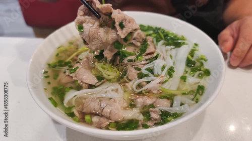 Delicious vietnamese pho bo for lunch on white table. Person eats vietnamese pho bo with chopsticks. This vietnamese pho bo is authentic cuisine with rice noodles and sliced meat and greens.