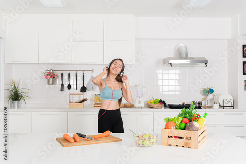 Cheerful young woman in blue sportswear enjoy listening to music on headphone while preparing fresh vegetable for a healthy meal in white kitchen.