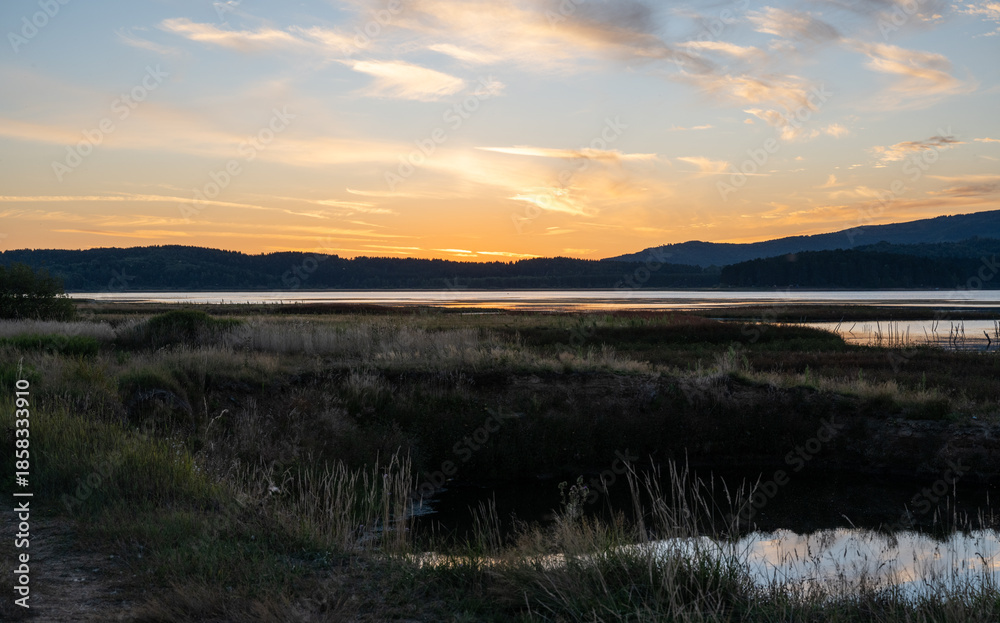 Obraz premium Minimalist dusk landscape featuring tall dry meadow grass, a reflective lake, and distant forested hills. 