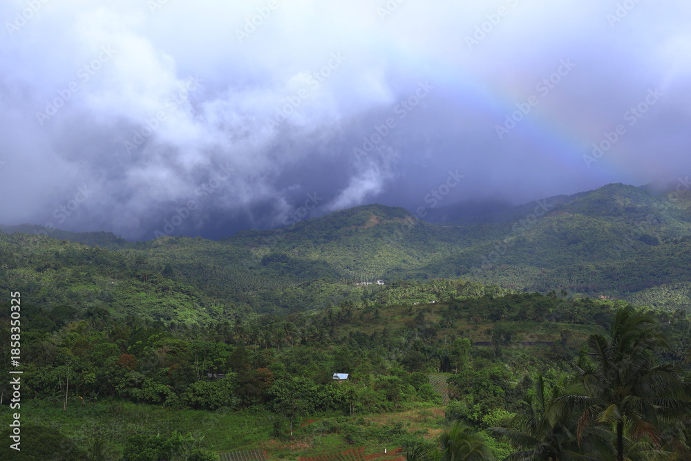 Naklejka premium morning in the mountains. The image is a scenic photograph of lush, green mountains in a tropical location, likely within the Philippines, with a rainbow visible in the upper right. 