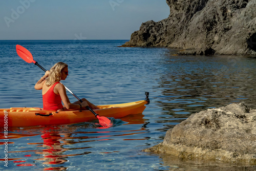 Kayaking, woman, sea, blonde woman paddles orange kayak on clear blue ocean near rocky coastline