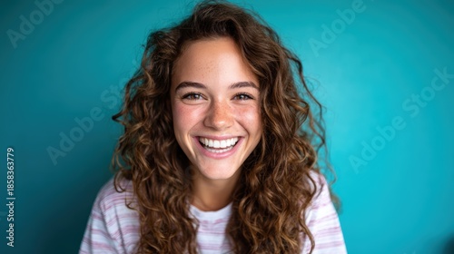 A cheerful young woman with curly hair beams joyfully against a vibrant turquoise background, exuding positivity and warmth that invites viewers to share in her happiness.