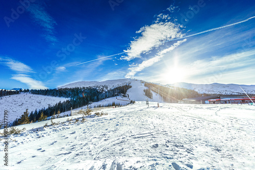 A wide scenic shot of a groomed ski slope in the Low Tatras, Slovakia, under a dramatic cloudy sky during twilight, featuring snow-covered ridges and evergreen forests.