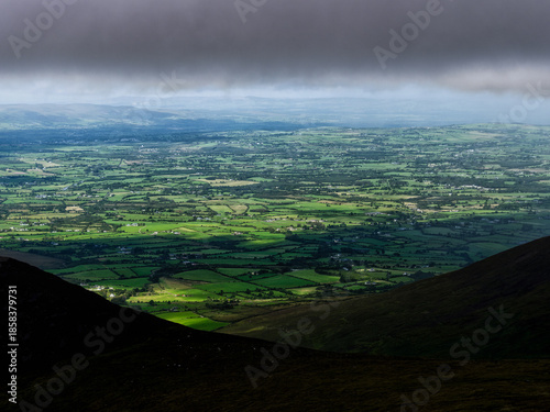 From a high vantage point on a mountain, one can see farmland, green fields, and small towns in Ireland. Dark clouds loom over the landscape.
