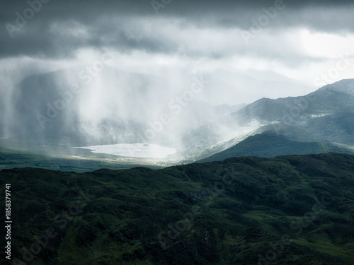 Lake surrounded by rolling hills with sunlight breaking through dark clouds and rain shafts.