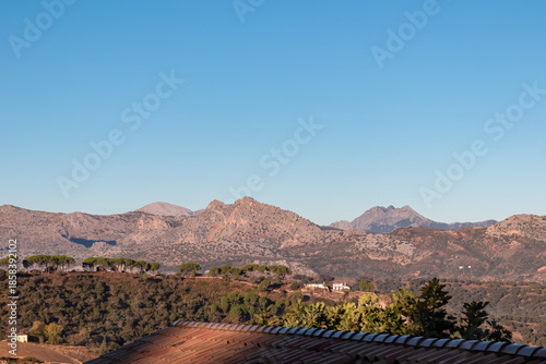 A terracotta roof frames green trees and rugged mountain peaks standing against a clear blue sky in the rural landscape of Ronda, Spain, capturing the scenic beauty of the vast Andalusian countryside.