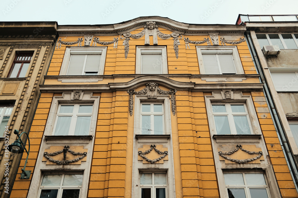 Obraz premium Yellow historic building facade with ornate stucco details and tall windows in morning Sarajevo. Classical architecture reflects urban heritage and calm atmosphere of the old city.