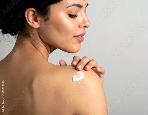 Woman with bare shoulder applying cream, looking elegant against a soft gray background