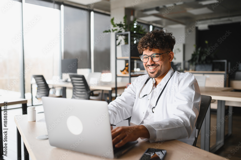 Fototapeta premium Smiling arab male doctor working on laptop in modern clinic office