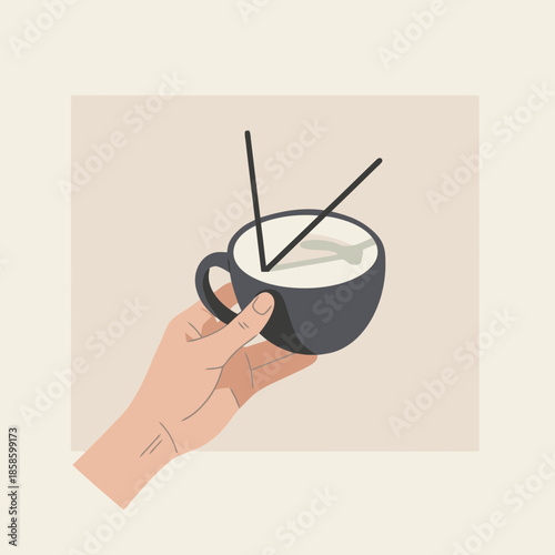 A hand holding a dark mug filled with a frothy white beverage and two chopsticks resting on top.
