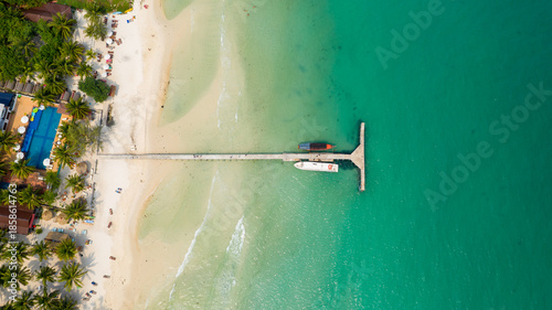 Aerial perspective shows a concrete pier extending from white sand into clear turquoise water at Coconut Beach, Koh Rong, Cambodia. Palm trees, a swimming pool, sun loungers, and anchored boats create