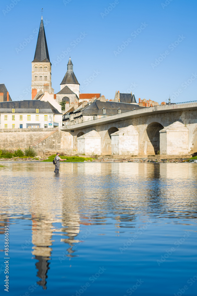 Fototapeta premium A solitary fisherman stands in the shallow waters of the Loire river, with the historic stone bridge and abbey spire of La Charite sur Loire rising above the riverside town. Clear blue sky and calm