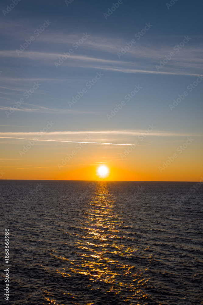 Fototapeta premium Golden sunlight stretches across calm open water as the sun sets above the horizon, creating a luminous path between the Adriatic and Ionian Seas. Wispy clouds and a gradient sky enhance the serene