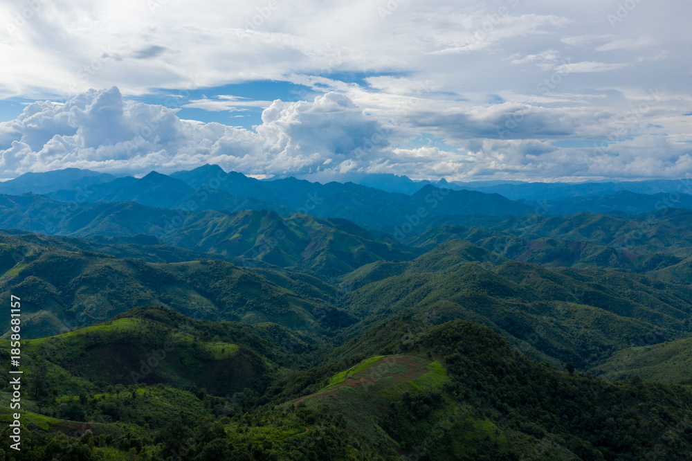 Obraz premium Vast mountain ridges covered in dense green vegetation stretch into the distance beneath a sky filled with towering cumulus clouds in the rural region between Phonsavan and Phou Khoun, Laos. The