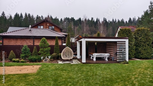 A wooden gazebo with a table, benches and a metal roof and a pine tree are decorated with glowing garlands for Christmas and New Year. There are houses, trees, lawn and swings nearby. Snowless winter