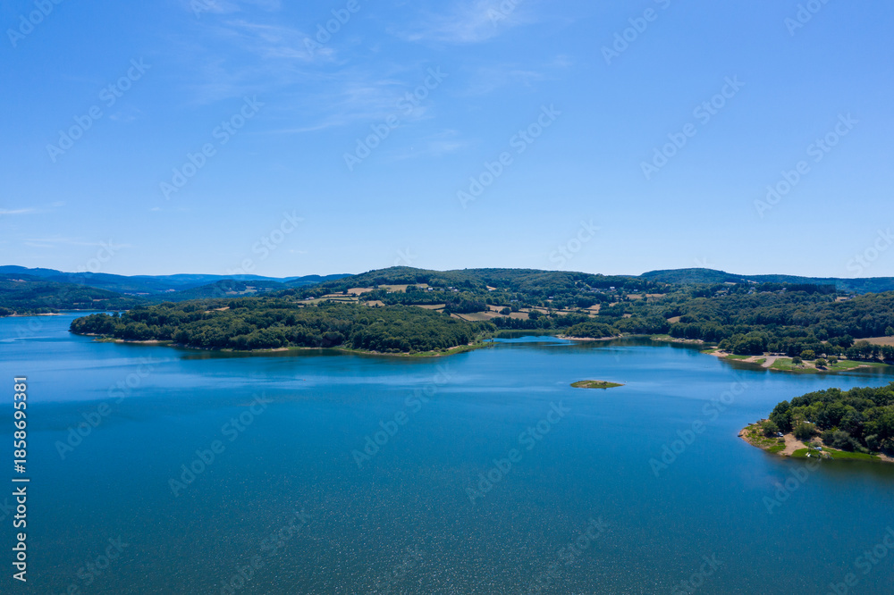 Obraz premium Wide view of Panneciere-Chaumard reservoir with deep blue water, surrounded by rolling forested hills and scattered farmland under a clear summer sky in Burgundy. The composition highlights tranquil
