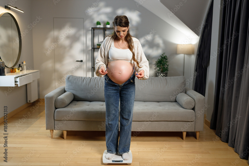 Fototapeta premium Expecting mother stands on scale in living room, checking weight gain during pregnancy, feeling clothes tightness