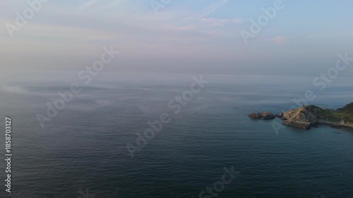 An aerial view of a tranquil seascape featuring calm, misty waters stretching to a hazy horizon and a small, rugged rocky island with vegetation.