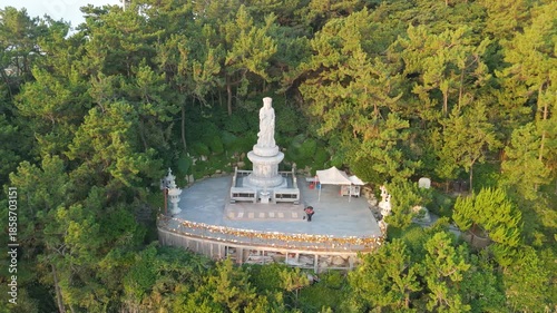 Aerial drone shot showcasing a majestic white marble statue of a Buddhist goddess on a high platform, surrounded by lush green trees and dense foliage, adorned with colorful offerings and traditional 