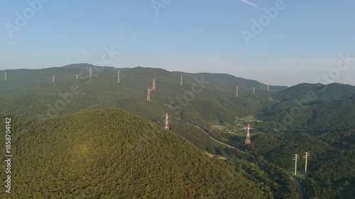 Lush green mountains dotted with numerous electricity transmission pylons under a clear blue sky, illustrating the coexistence of natural landscapes and essential energy infrastructure.