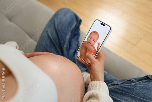Pregnant woman in denim jeans and white top holding smartphone displaying ultrasound image of baby while resting on sofa