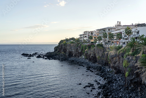 Coastal town on volcanic cliffs overlooking the Mediterranean Sea in Sicily, Italy. Scenic seaside cityscape with rocky shoreline, palm trees, residential buildings, and calm evening light