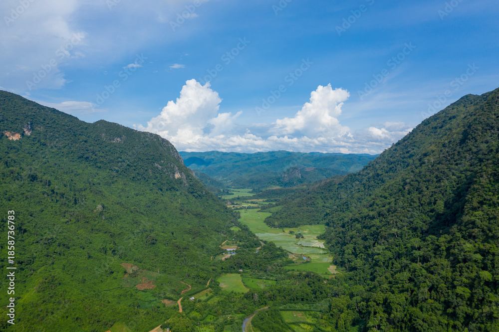 Naklejka premium Wide aerial view of a vibrant green valley surrounded by steep forested hills in northern Laos. The landscape features patchwork rice fields, dense jungle, and a bright blue sky with dramatic cloud