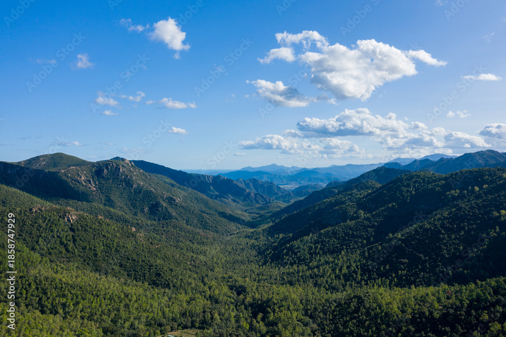 Fototapeta premium Aerial view of a vast green valley surrounded by rugged mountains and dense woodland in Sardinia, Italy. Puffy clouds drift across a clear blue sky, casting gentle shadows over the landscape.