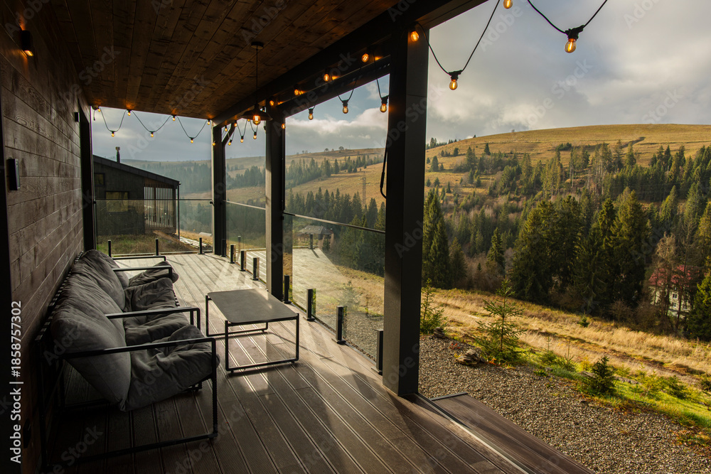 Fototapeta premium Balcony with seating and table, with lights hanging above, view showing rolling hills and trees under a cloudy sky in the mountains