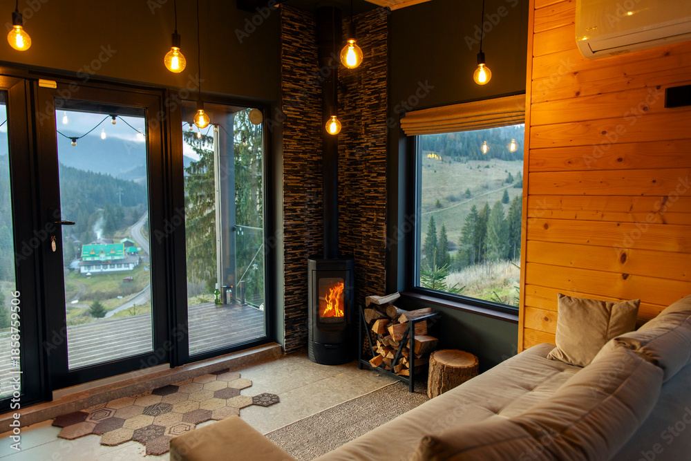 Naklejka premium Cozy living room with panoramic window and mountains view, wood stove in the corner, comfy sofa and lights hanging from the ceiling