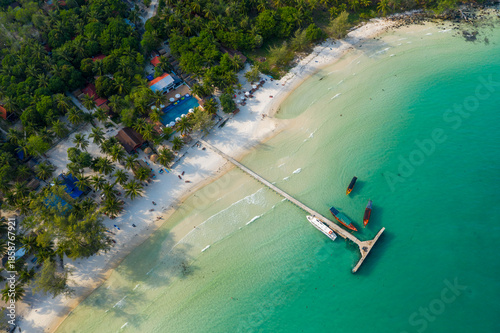 Overhead perspective shows a long wooden pier stretching from white sand into clear turquoise sea at Coconut Beach on Koh Rong island, Cambodia. Palm trees, scattered boats, and lush tropical