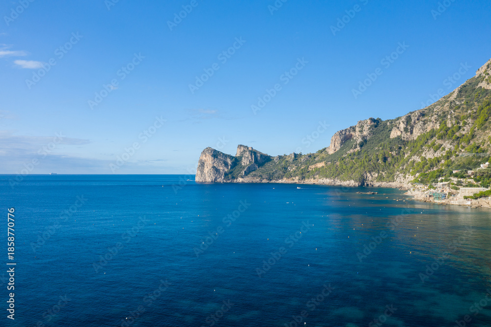 Fototapeta premium Clear blue waters meet rugged limestone cliffs along a tranquil stretch of the Italian countryside coastline. Sunlight highlights the lush greenery and rocky shoreline under a vivid sky, evoking a