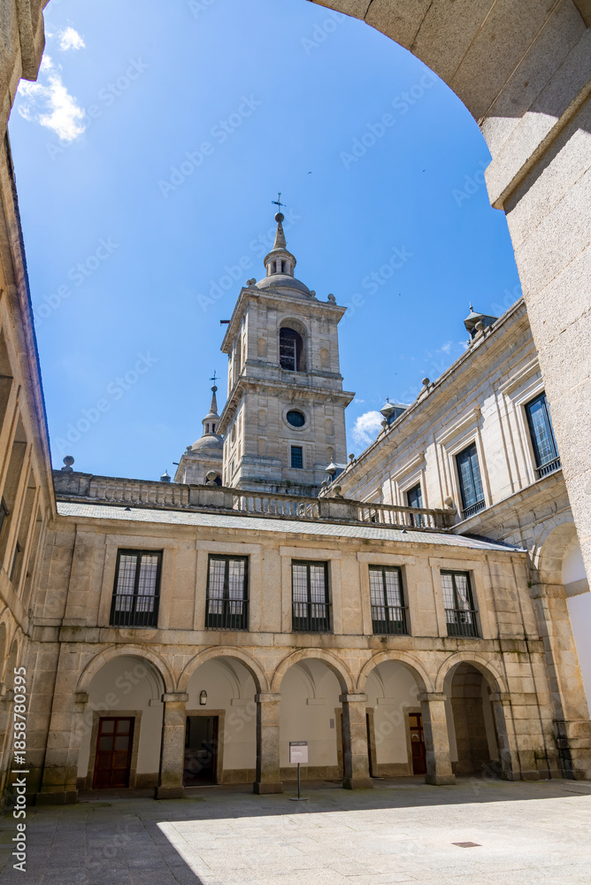 Obraz premium A sunny view of a symmetrical inner patio at El Escorial in Spain, featuring massive granite towers, uniform facades, and a grand basilica entrance. San Lorenzo
