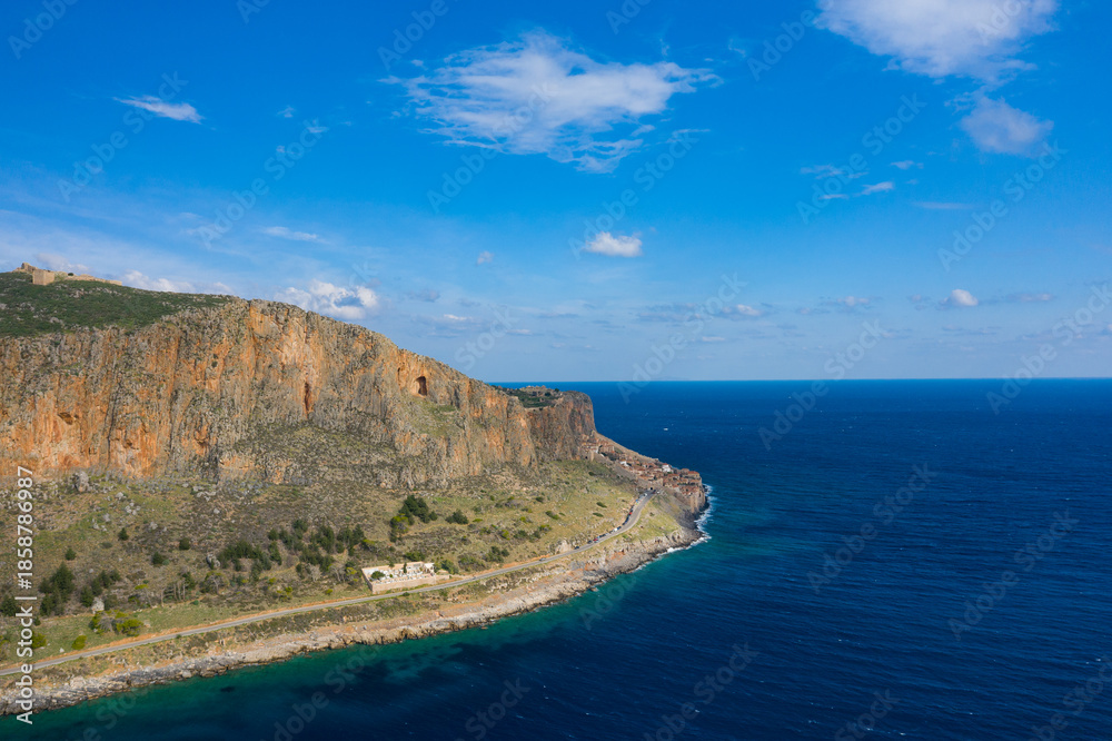 Obraz premium Steep orange cliffs of Monemvasia overlook a winding coastal road and vivid turquoise sea beneath a wide blue sky. The dramatic landscape is bathed in sunlight, highlighting rocky textures and natural