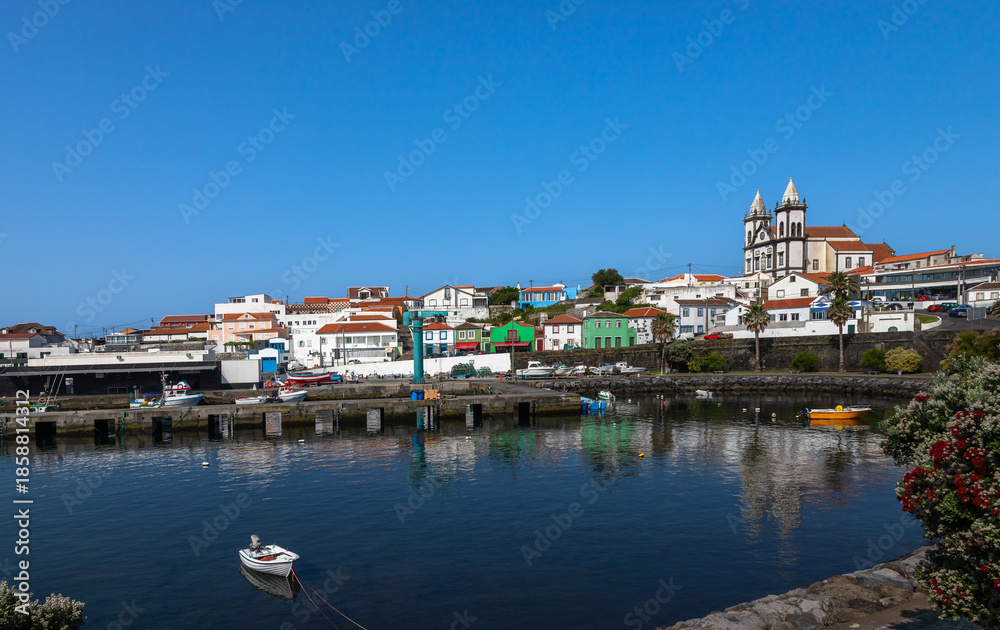 Fototapeta premium Scenic view of São Mateus da Calheta harbor on Terceira Island, Azores, Portugal, featuring colorful houses, fishing boats, calm waters, and a historic church under a clear blue sky