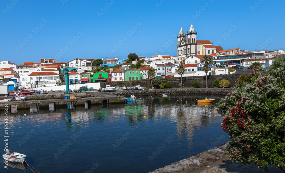 Fototapeta premium Scenic view of São Mateus da Calheta harbor on Terceira Island, Azores, Portugal, featuring colorful houses, fishing boats, calm waters, and a historic church under a clear blue sky