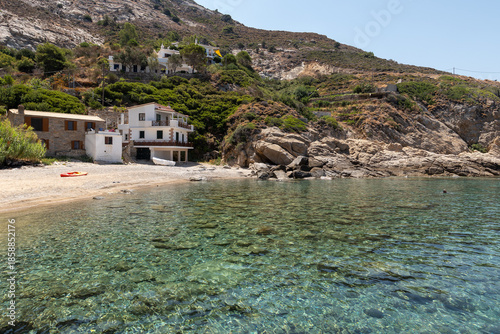 Clear transparent water along Nanouras beach with traditional houses in the background on Ikaria island, Greece. Mediterranean seaside village and summer travel destination.