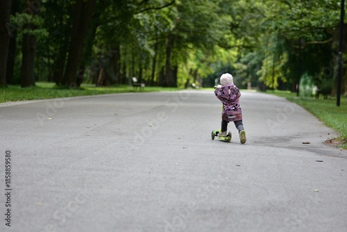 Wallpaper Mural Toddler riding a three-wheel scooter along a park path, back view. Child enjoying freedom, movement and outdoor activity surrounded by green trees and peaceful nature. Torontodigital.ca
