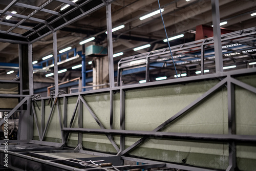 Industrial vehicle manufacturing workshop with metal bus frames and body structures under assembly. Large factory interior shows fabrication, welding, and custom vehicle production in progress.