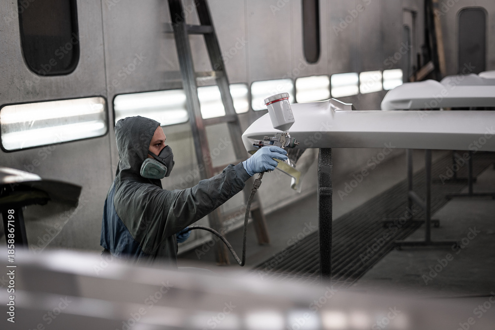 Fototapeta premium Worker spray painting vehicle body part in professional paint booth. Industrial automotive painting process with protective equipment, precision tools, and controlled environment.