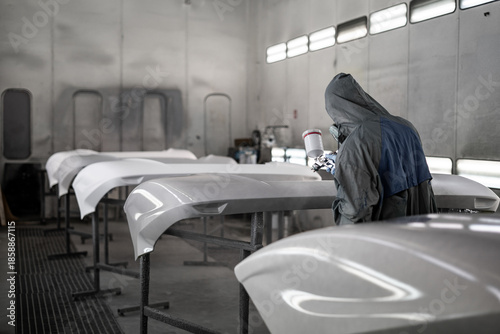 Worker spray painting vehicle body part in professional paint booth. Industrial automotive painting process with protective equipment, precision tools, and controlled environment.