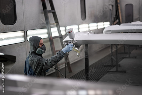 Worker spray painting vehicle body part in professional paint booth. Industrial automotive painting process with protective equipment, precision tools, and controlled environment.