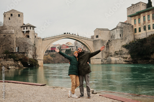 Photos Smiling couple with open arms standing by the river near the historic bridge in Mostar